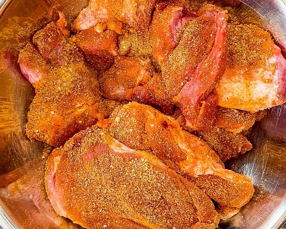 Pork shoulder coated with dry rub seasoning in a bowl before braising in a Dutch oven.