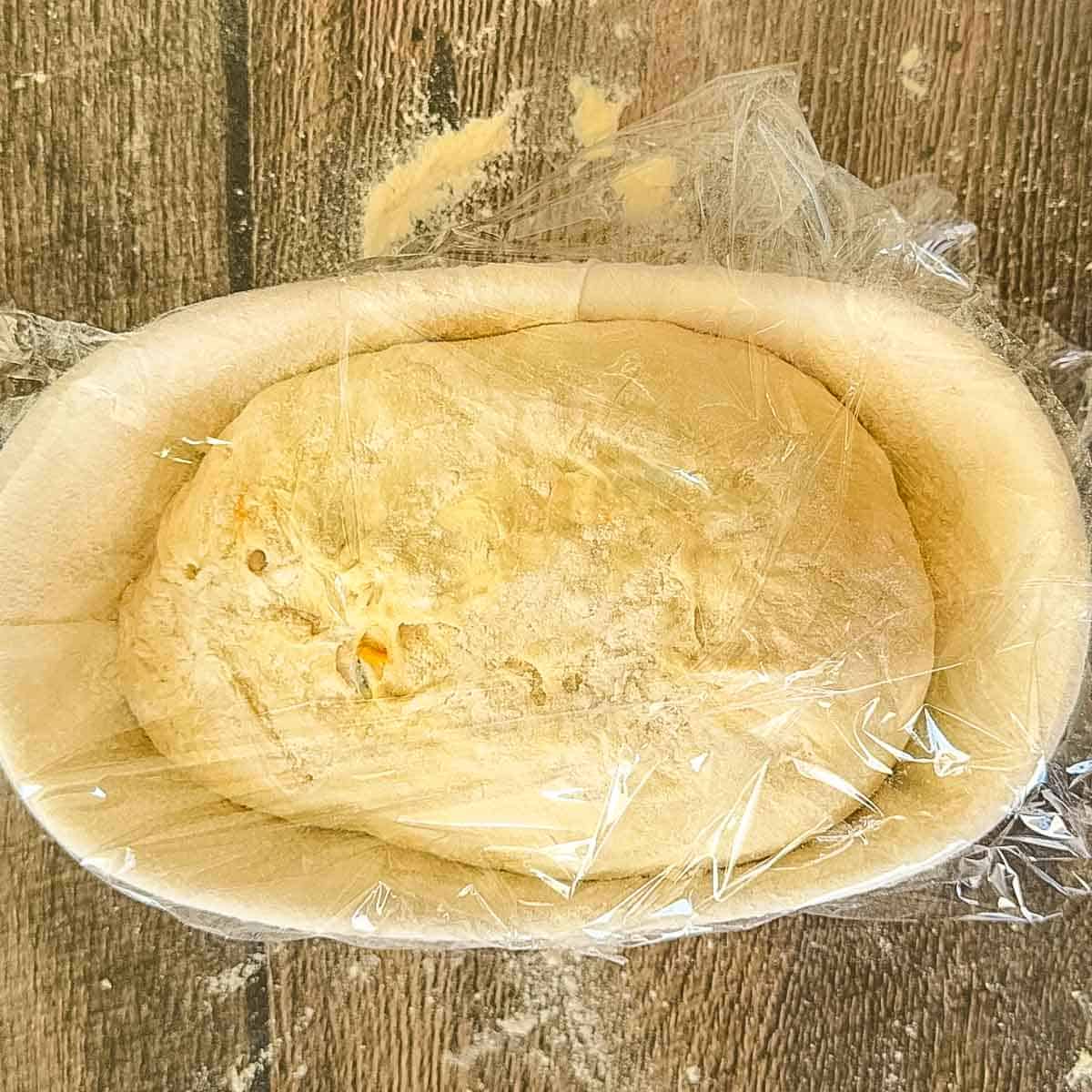 The shaped cheddar jalapeno dough in a floured banneton basket, covered with plastic.