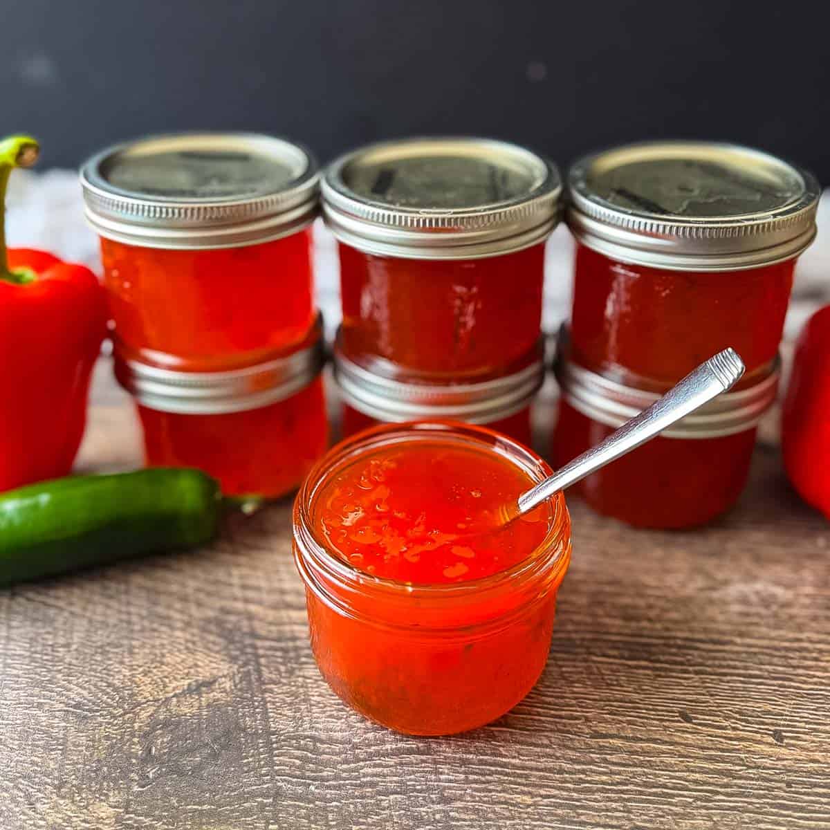 Homemade red pepper jelly made with chopped peppers, shown in an open jar with sealed jars behind.