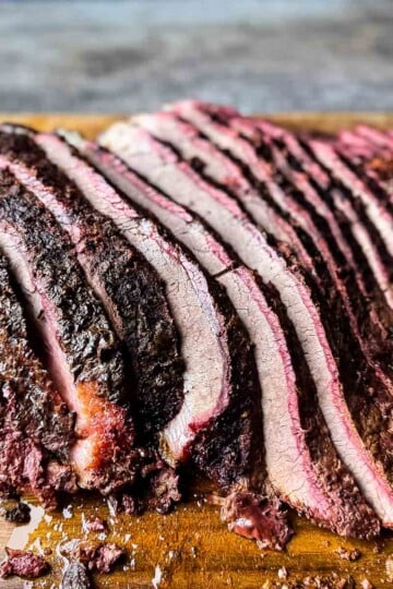 Sliced hot and fast smoked brisket with a dark bark and visible smoke ring resting on a wooden cutting board.