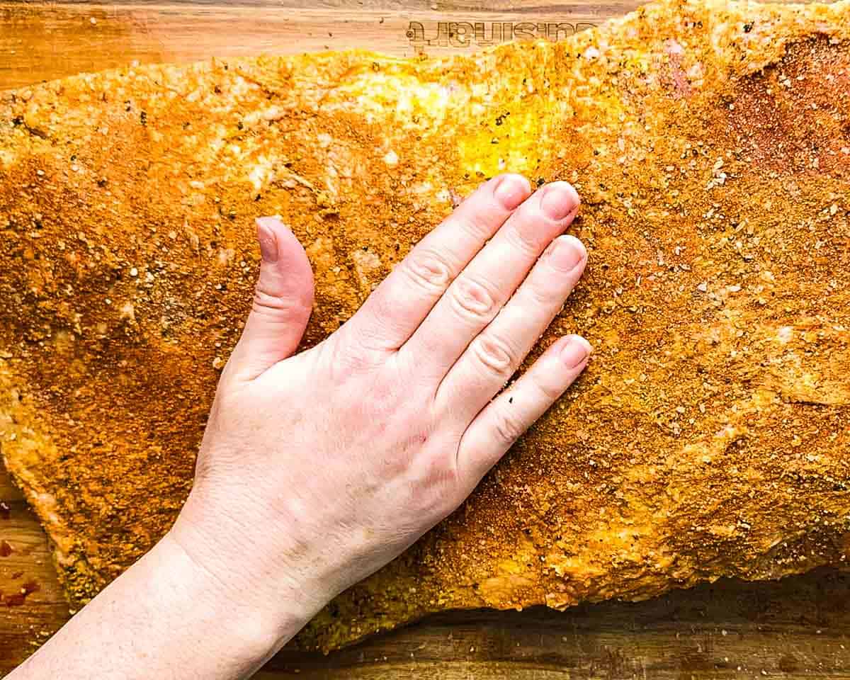 Pressing dry rub onto a mustard-coated brisket before smoking.