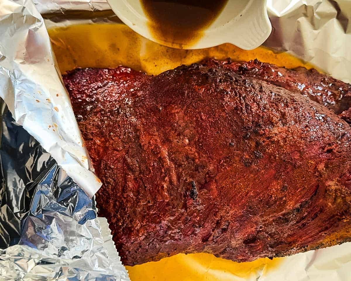 The braising liquid being poured beside the brisket before it gets wrapped in foil.