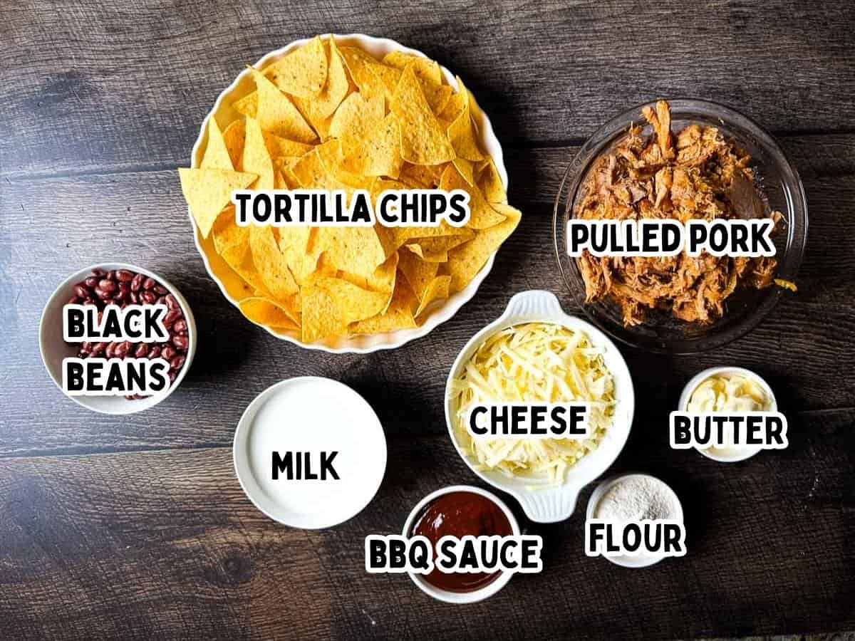 Labeled ingredients for BBQ pulled pork nachos in individual bowls on a wooden table.