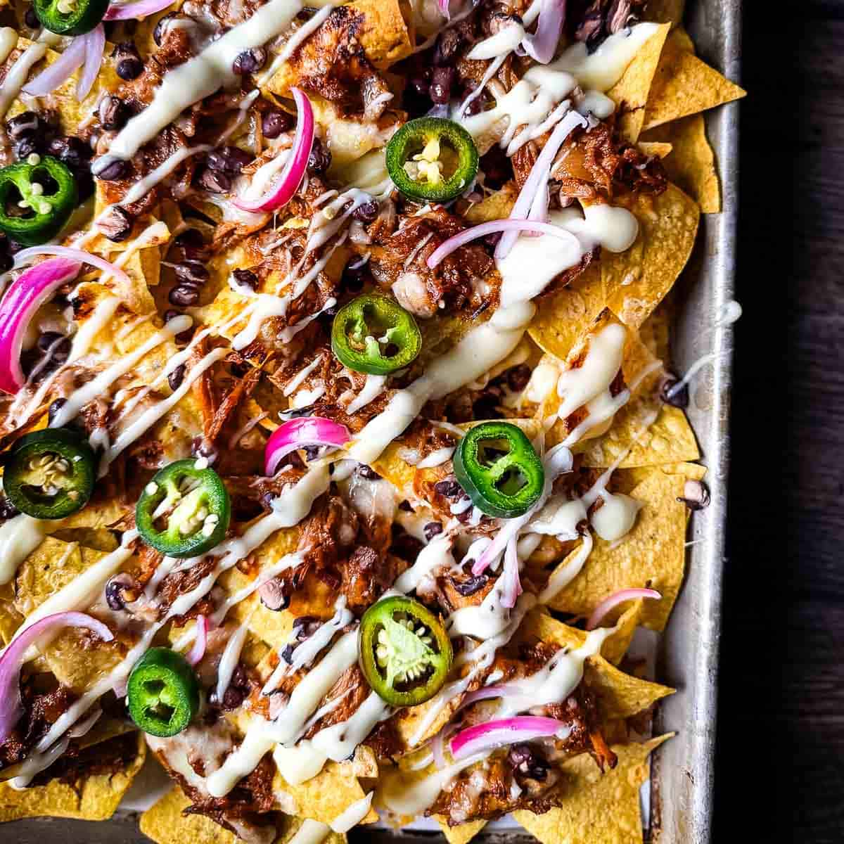 Loaded pulled pork nachos on a baking sheet with jalapeños, red onion, and melted cheese.