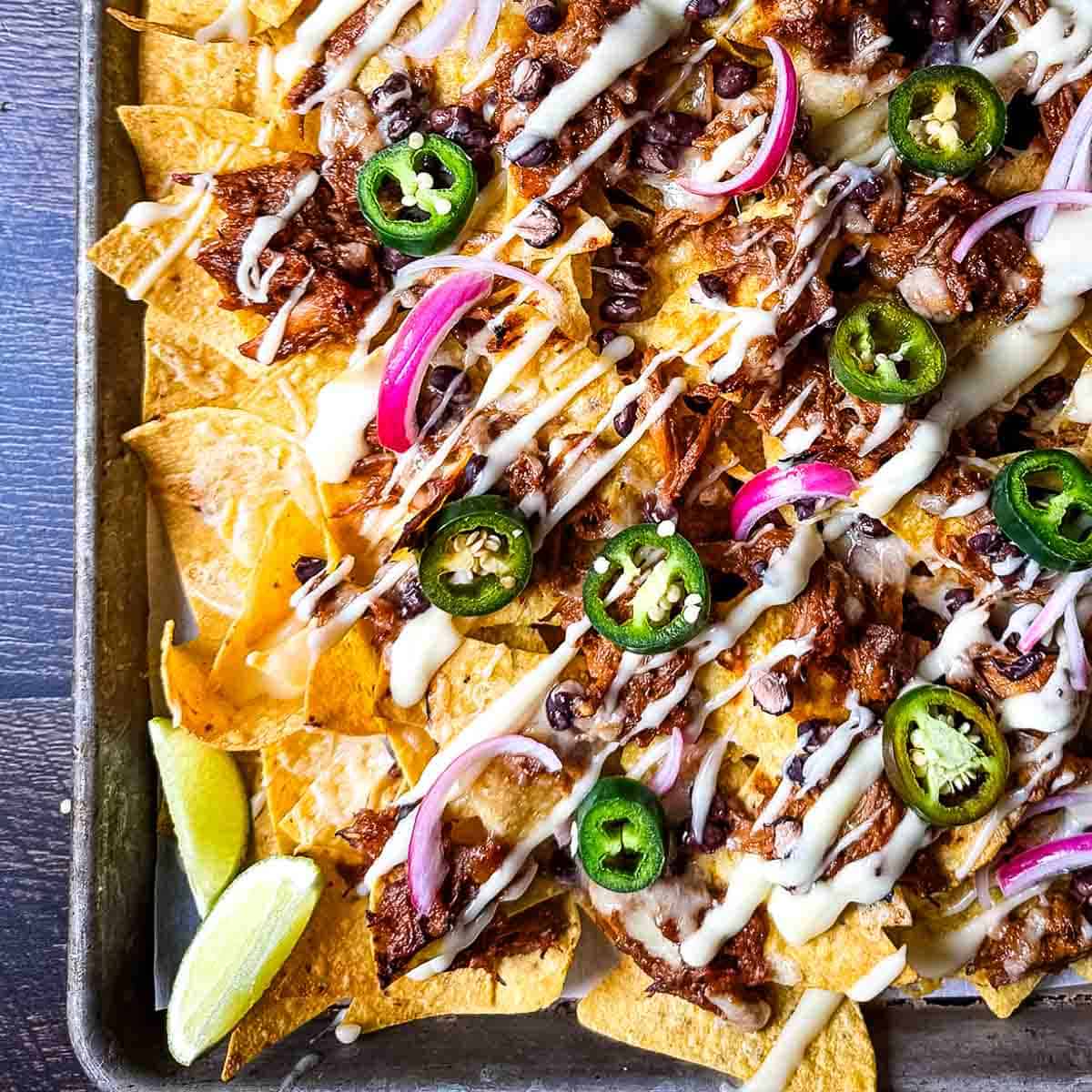 Sheet pan of loaded nachos topped with pulled pork, sliced jalapeños, red onion, black beans, and a creamy sauce drizzle.