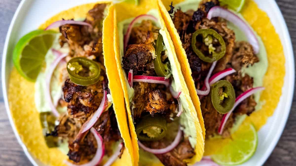 Plate of carnitas street tacos topped with crispy pork, pickled onions, jalapeños, and avocado crema served with fresh lime wedges.