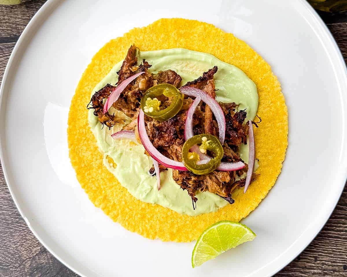 A corn tortilla topped with avocado crema, shredded crispy pork, pickled red onion, and pickled jalapeno.