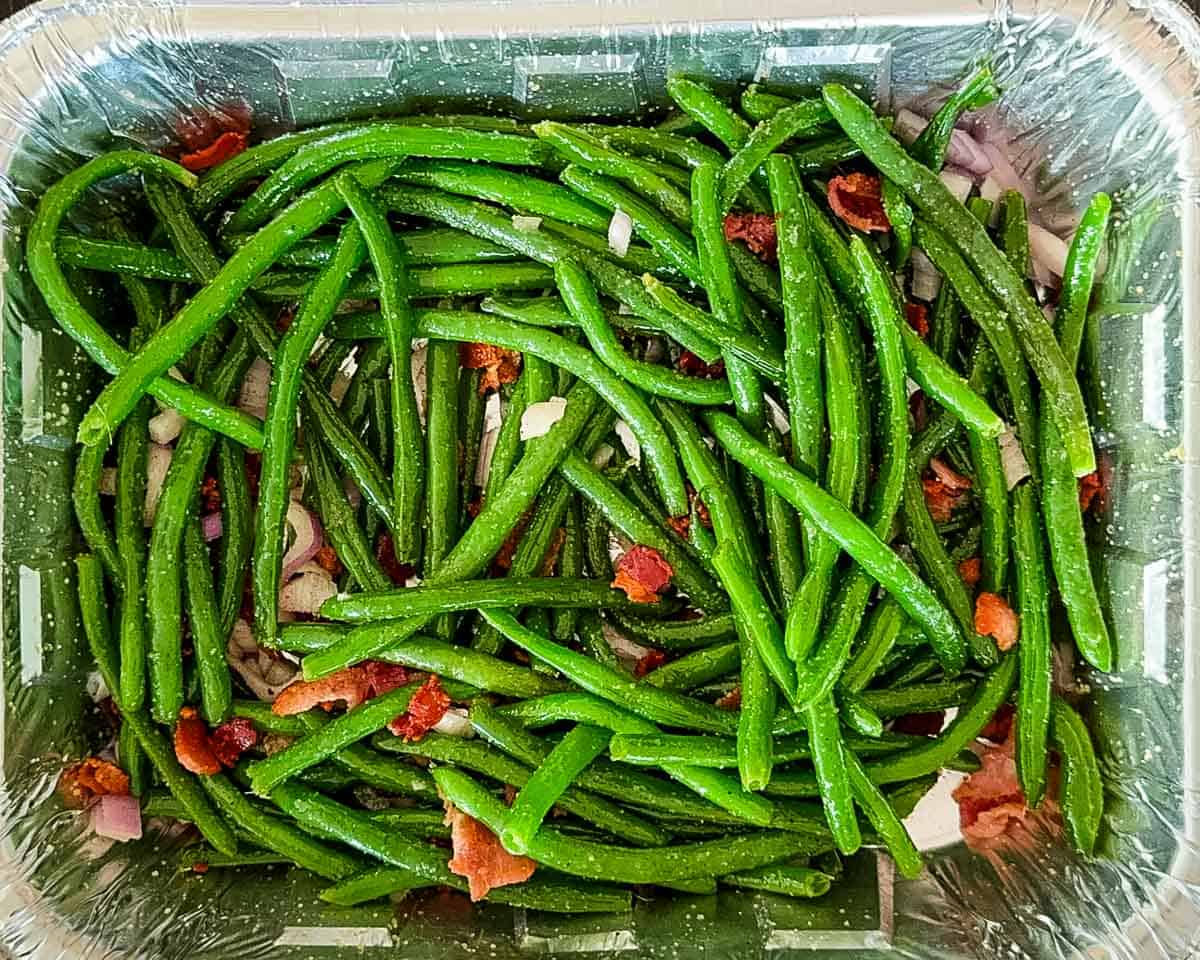 The green beans tossed in a foil pan with seasoning, red onion, and bacon.