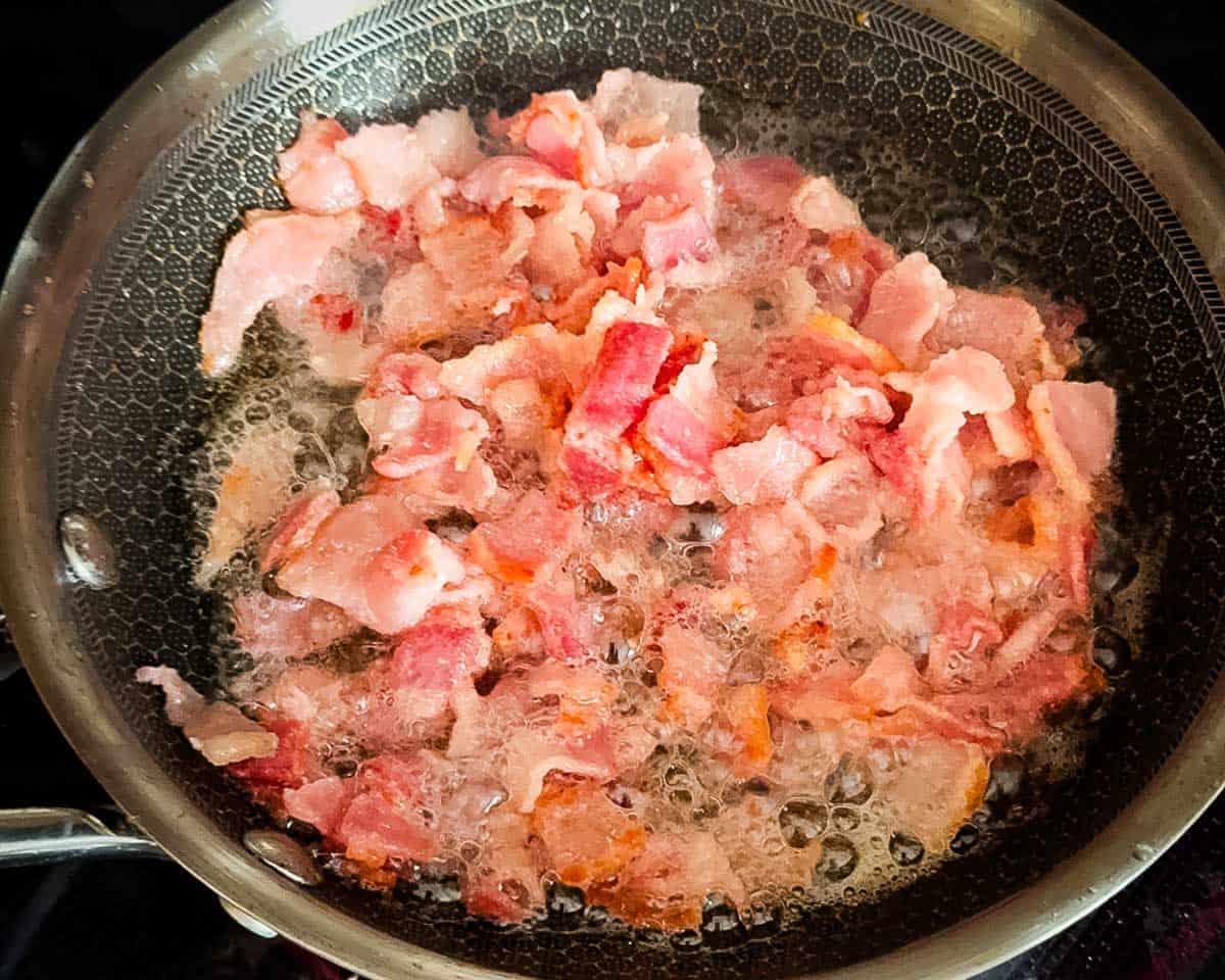 The bacon being cooked in a large skillet.