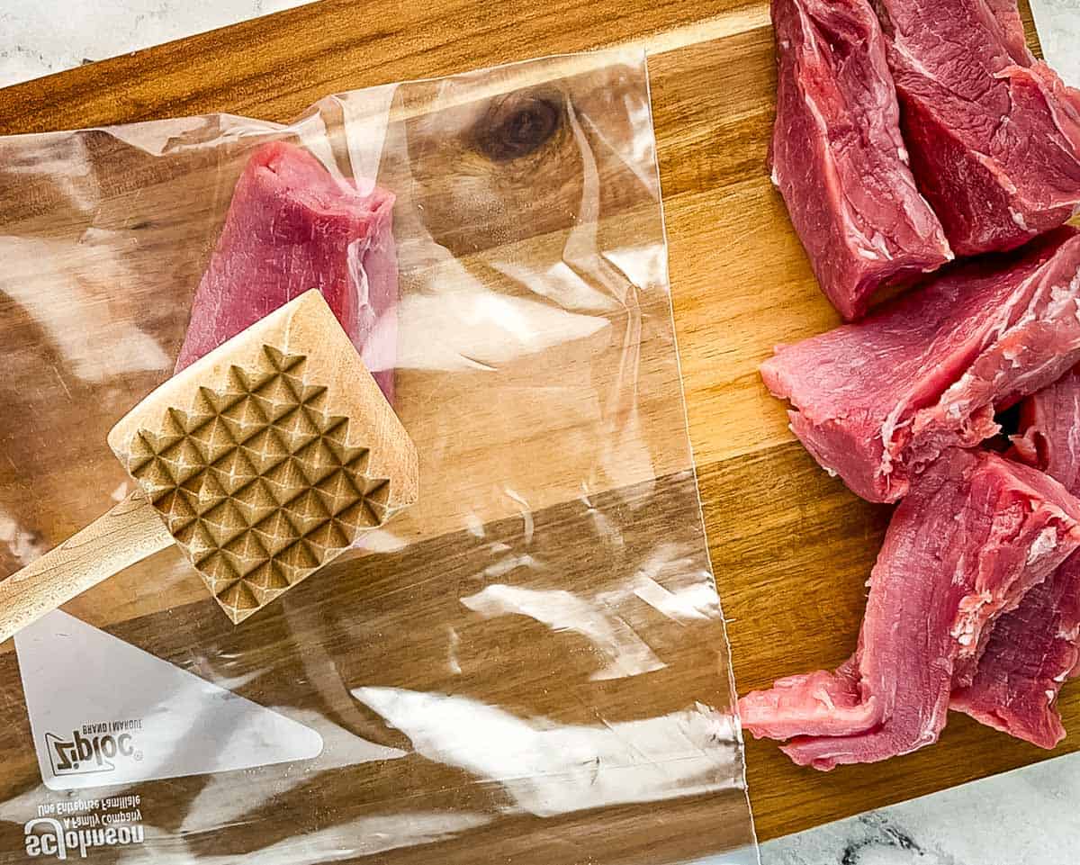 Pork pieces in a plastic bag being pounded with a meat mallet to an even thickness for air fryer pork tonkatsu.