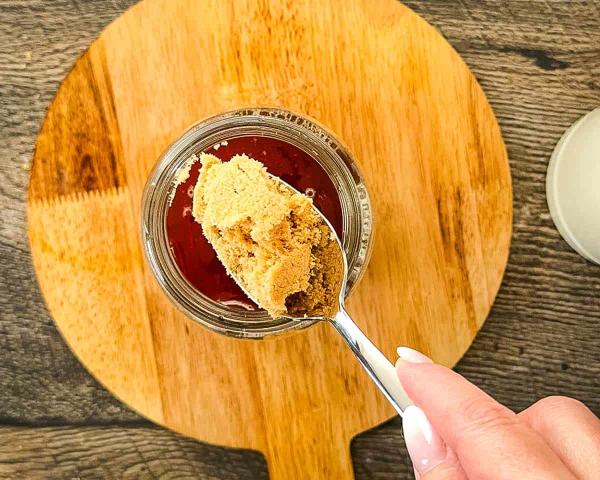 Adding brown sugar to warm chai concentrate in a glass jar before stirring.