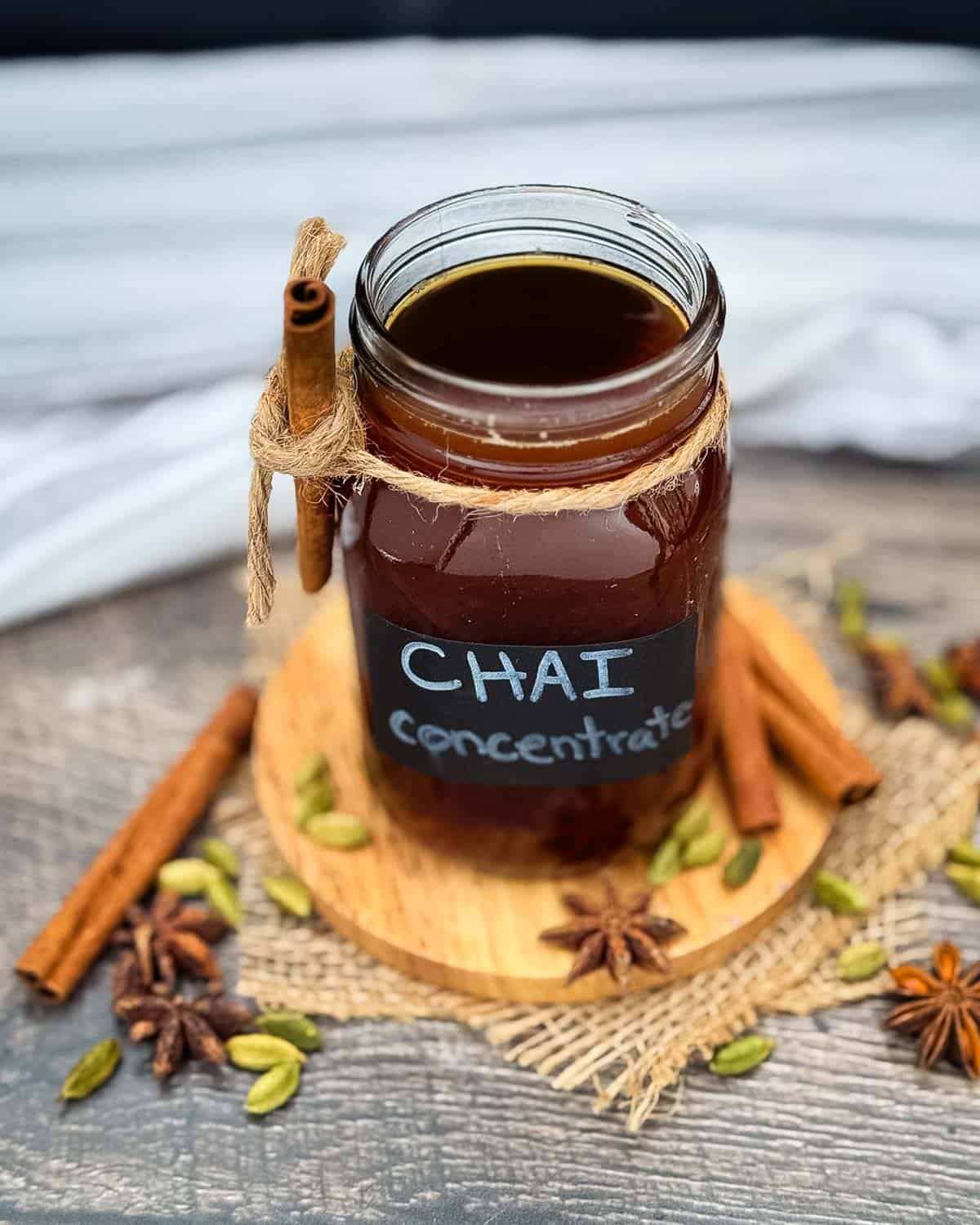 Glass jar of homemade chai tea concentrate surrounded by cinnamon sticks, star anise, and green cardamom pods.