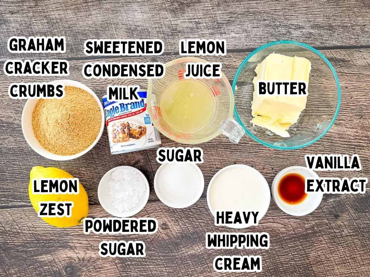 The ingredients for No-Bake Lemon Cream Pie arranged on a dark wood surface, including graham cracker crumbs, a can of sweetened condensed milk, fresh lemon juice, butter, a whole lemon for zest, powdered sugar, granulated sugar, heavy whipping cream, and vanilla extract.