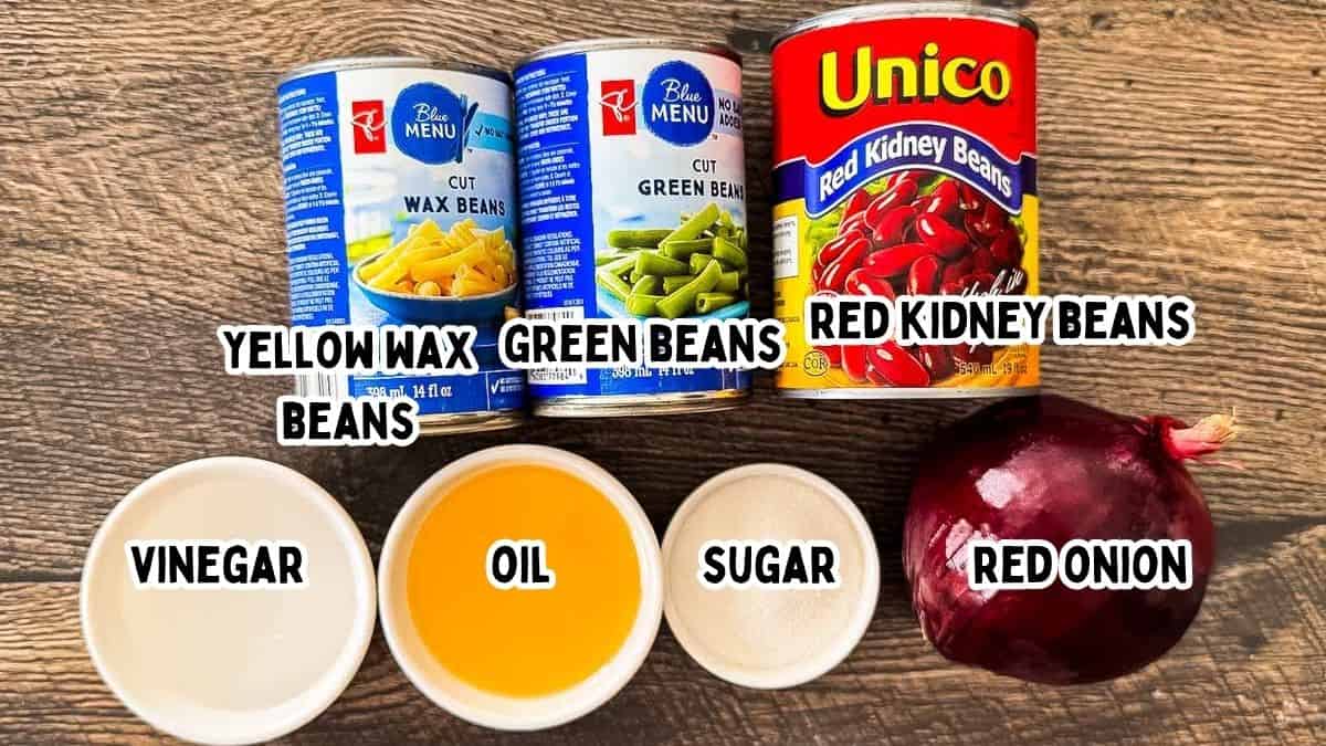 Labeled ingredients for three bean salad on a wooden table, including three kinds of beans, red onion, and vinegar, oil, and sugar for the dressing.