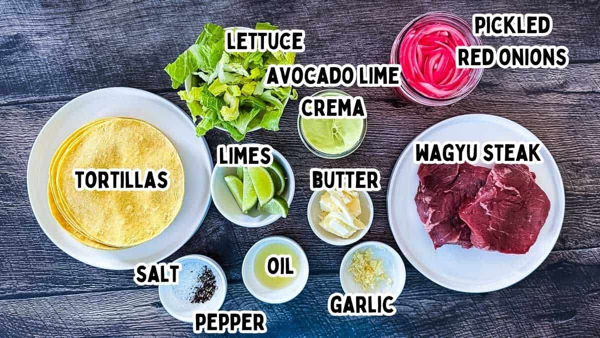 Ingredients for Wagyu steak tacos labeled with text including raw Wagyu steak, corn tortillas, lettuce, avocado lime crema, pickled red onions, butter, garlic, oil, limes, salt, and pepper.
