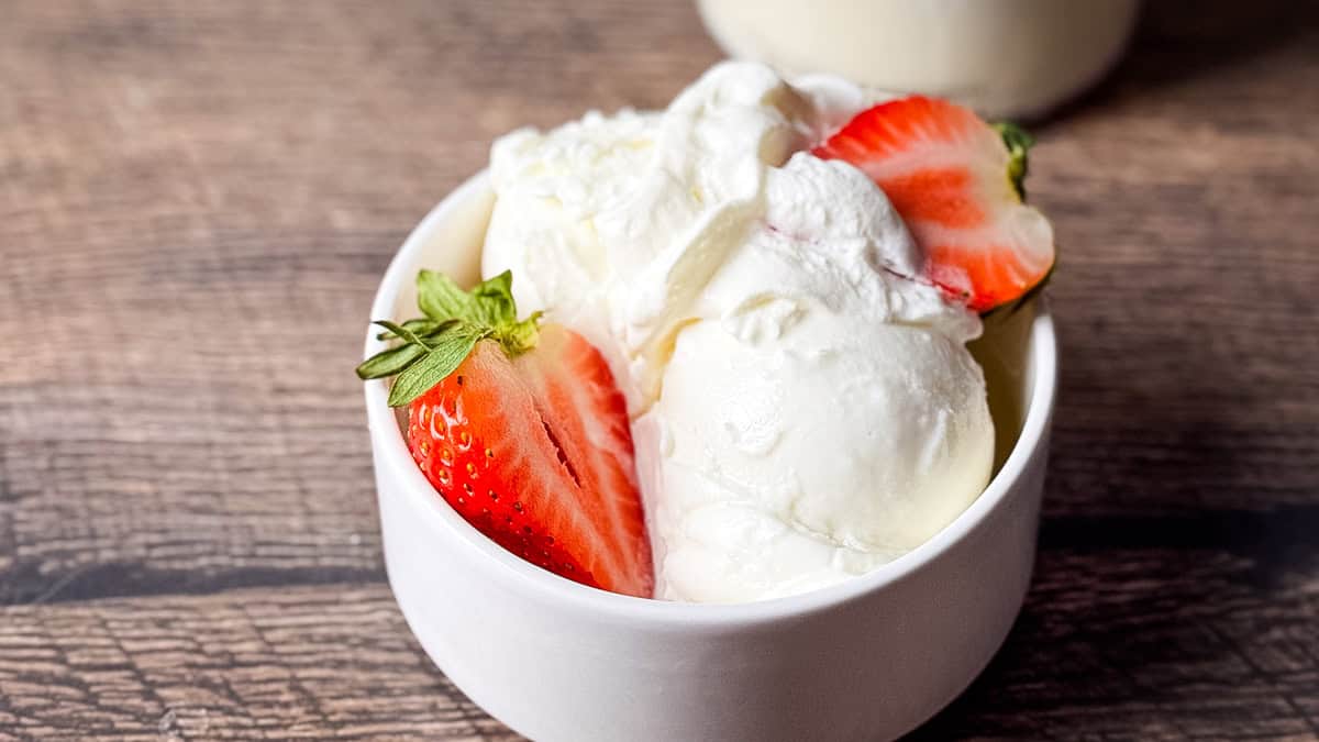 Rounded scoop of creamy Ninja Creami frozen vanilla yogurt in a white bowl, garnished with a fresh strawberry slice and whipped cream.