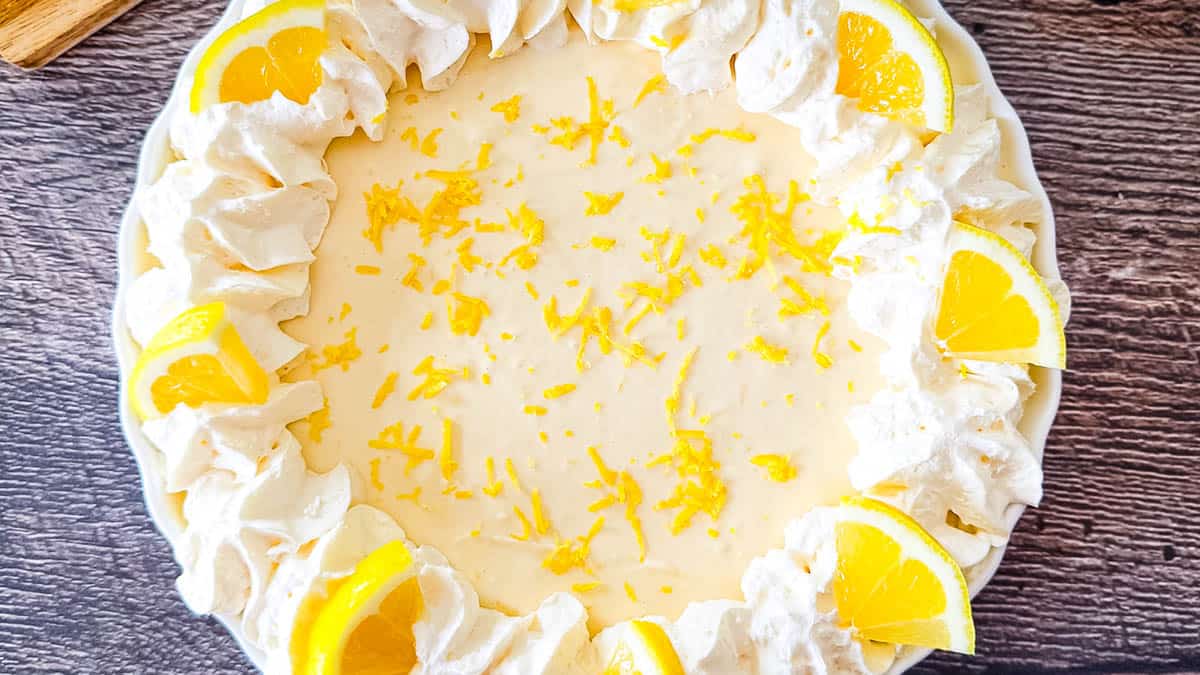 A top-down view of a finished No-Bake Lemon Cream Pie in a white scalloped pie dish, featuring a ring of piped whipped cream, fresh lemon slices, and bright yellow lemon zest.