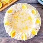 No-Bake Lemon Cream Pie in a white scalloped pie dish, featuring a ring of piped whipped cream, fresh lemon slices, and bright yellow lemon zest on a dark wood background.