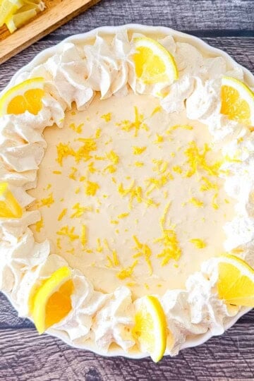 No-Bake Lemon Cream Pie in a white scalloped pie dish, featuring a ring of piped whipped cream, fresh lemon slices, and bright yellow lemon zest on a dark wood background.