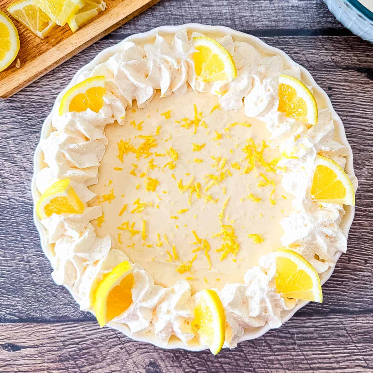 No-Bake Lemon Cream Pie in a white scalloped pie dish, featuring a ring of piped whipped cream, fresh lemon slices, and bright yellow lemon zest on a dark wood background.