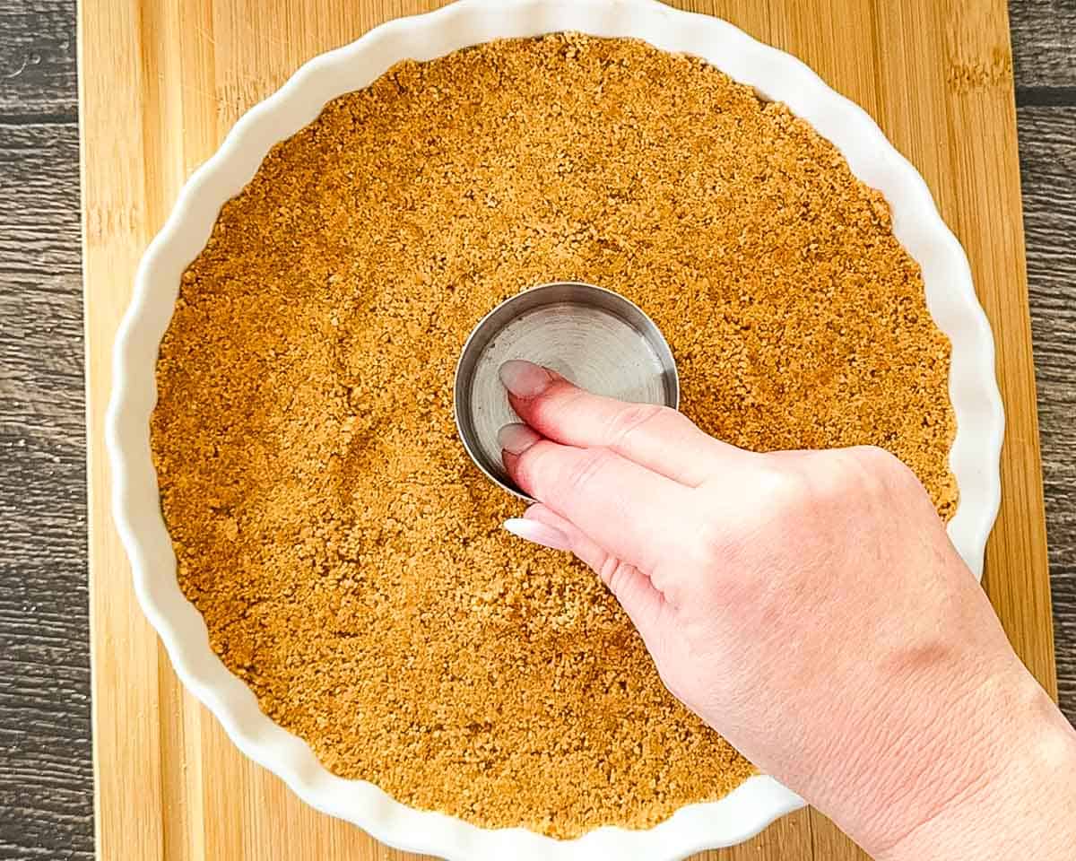 A graham cracker crust being firmly pressed into a white scalloped pie dish, ready for the lemon filling.