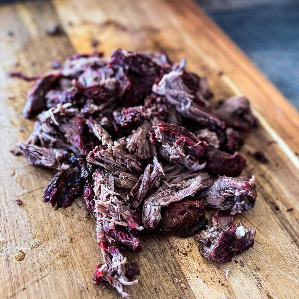 Smoked pulled beef made from chuck roast, shredded into juicy pieces with a dark bark on a wooden cutting board.