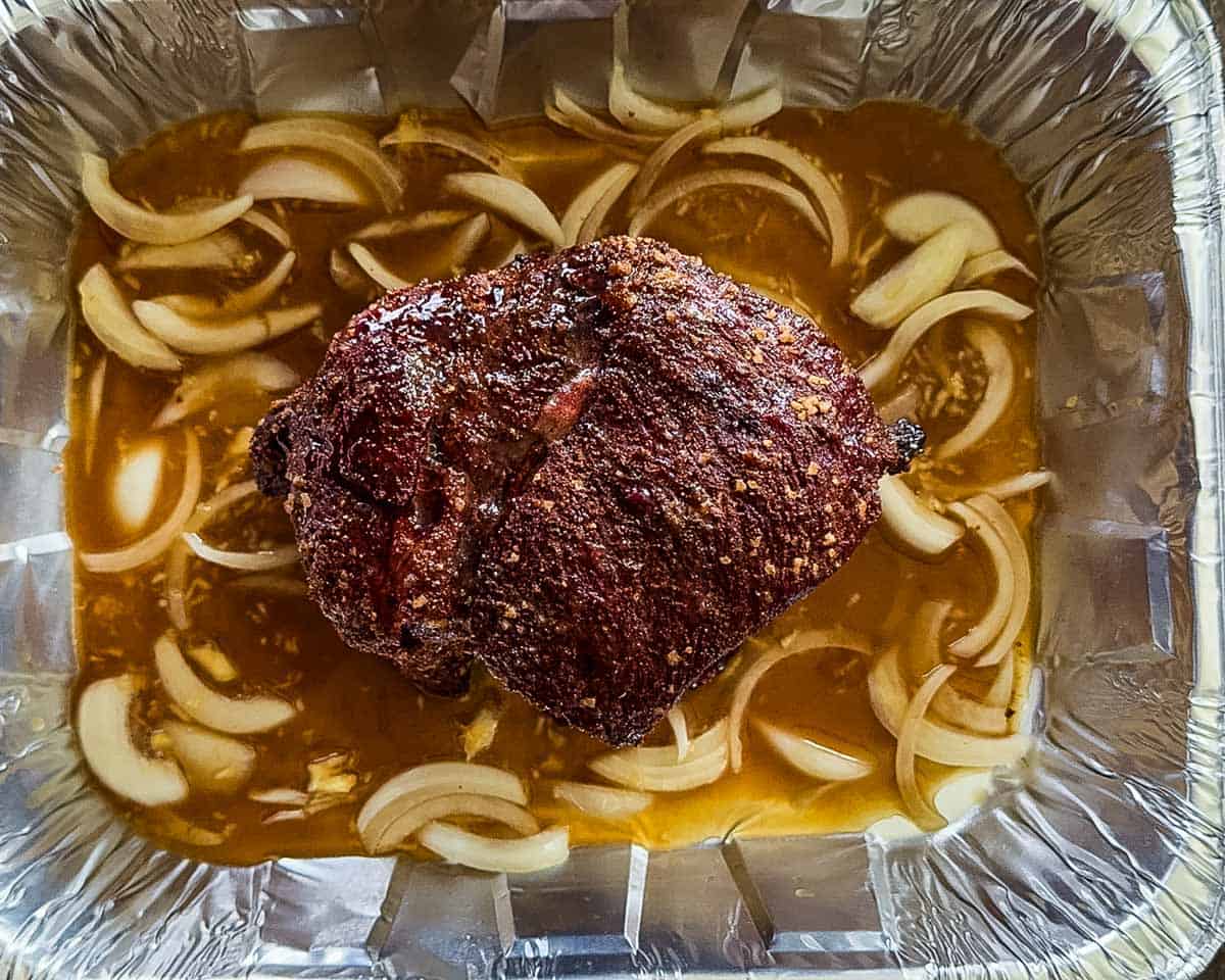 The smoked beef roast in a foil pan with onions, garlic, and broth.