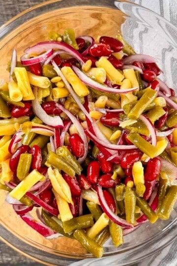 Three bean salad with kidney beans, green beans, yellow wax beans, and sliced red onion in a glass bowl.