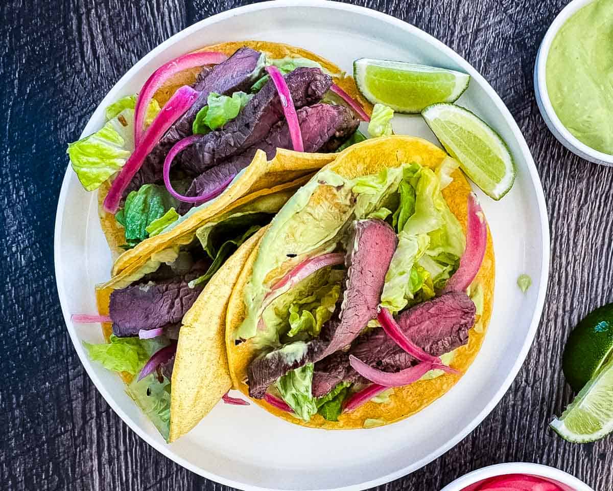 Three steak tacos on a white dinner plate, topped with avocado lime crema and pickled red onions.