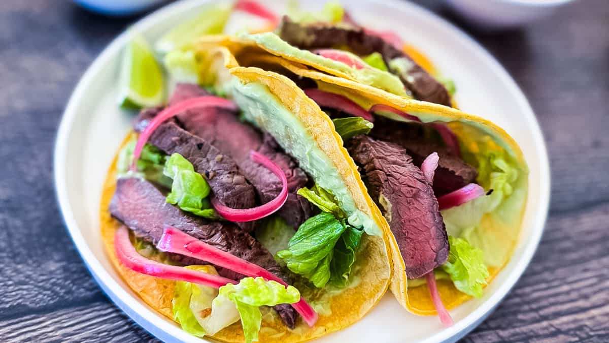 Close-up of three Wagyu steak tacos on corn tortillas with bright pink pickled red onions, fresh lettuce, and lime wedges on a white plate.