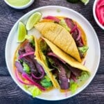 Two seared Wagyu steak tacos in corn tortillas topped with pickled red onions, shredded lettuce, and avocado lime crema, served on a white plate with lime wedges.