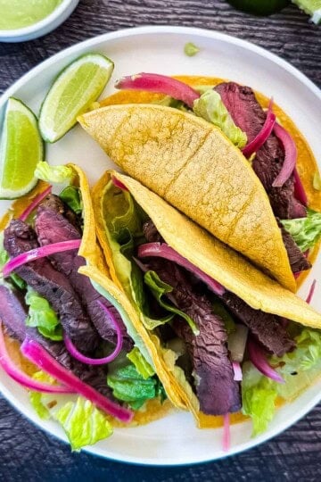 Two seared Wagyu steak tacos in corn tortillas topped with pickled red onions, shredded lettuce, and avocado lime crema, served on a white plate with lime wedges.