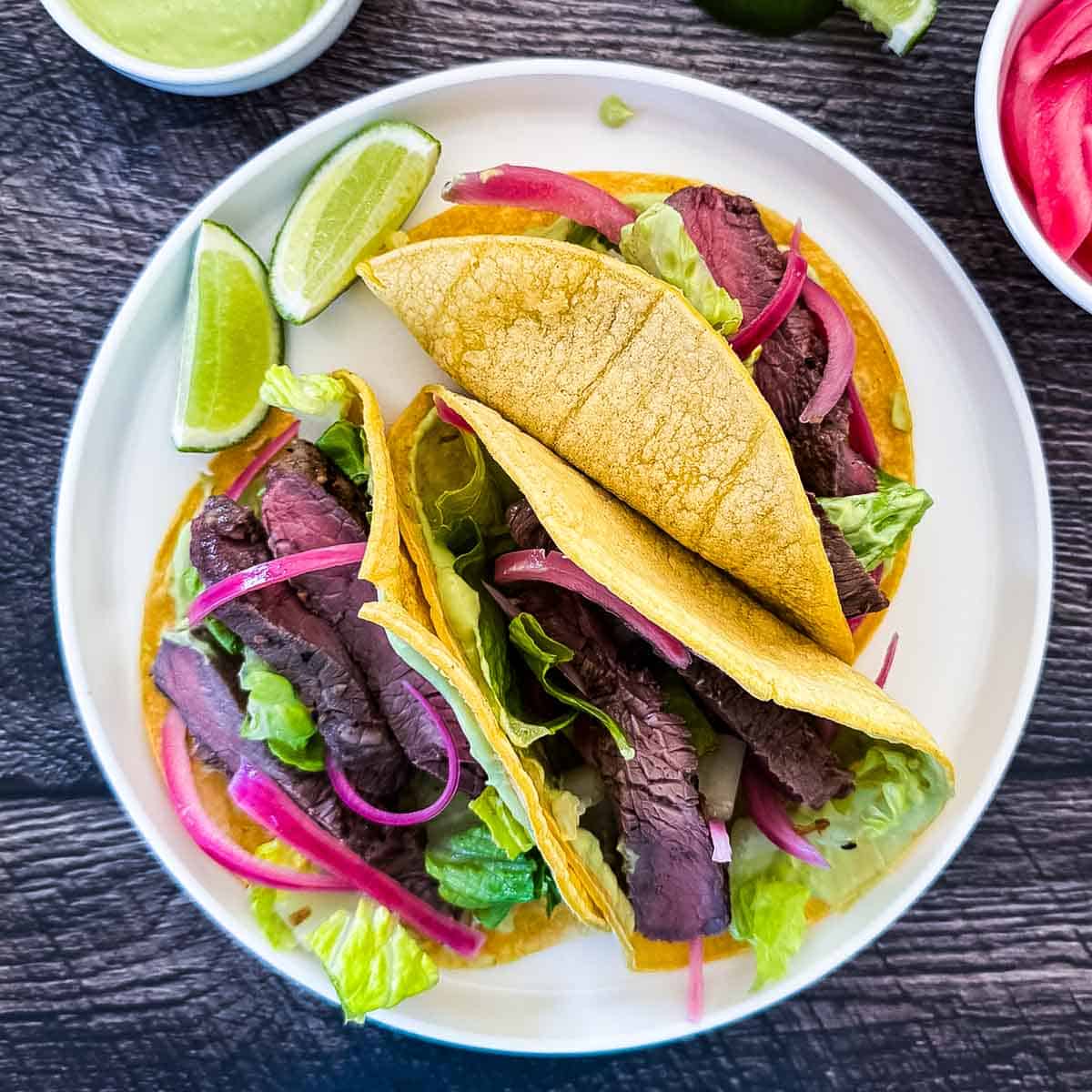 Two seared Wagyu steak tacos in corn tortillas topped with pickled red onions, shredded lettuce, and avocado lime crema, served on a white plate with lime wedges.