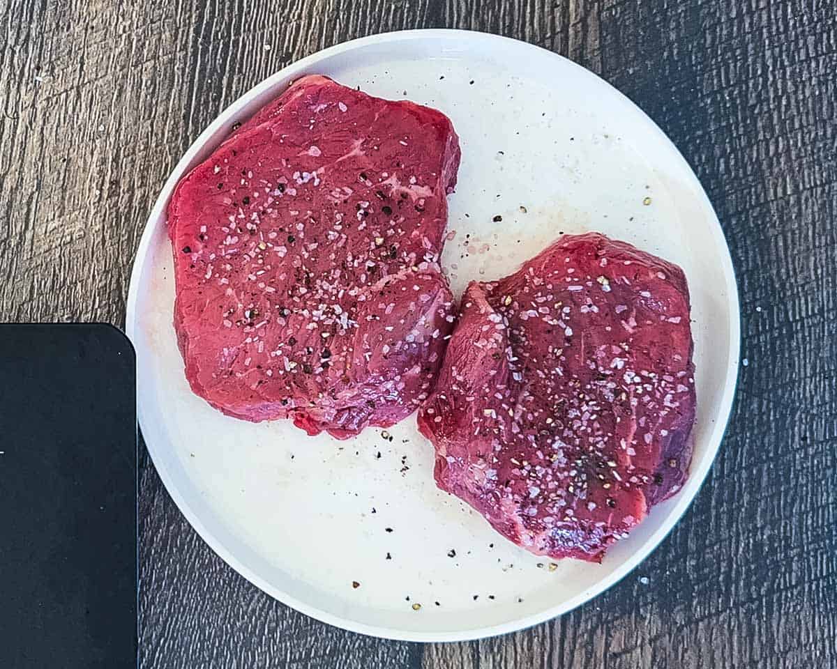 Oiled and seasoned steaks on a white plate.