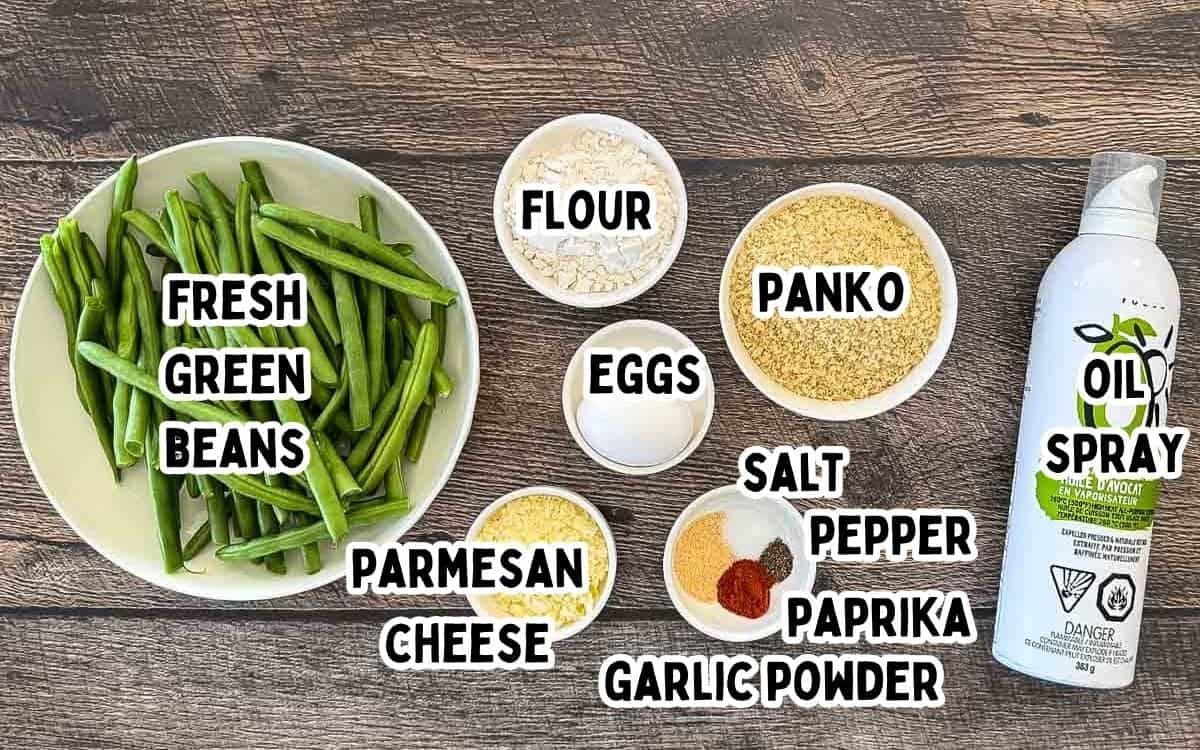 Fresh green beans, flour, eggs, panko breadcrumbs, Parmesan cheese, garlic powder, paprika, salt, black pepper, and oil spray for air fryer green bean fries on a wooden table.