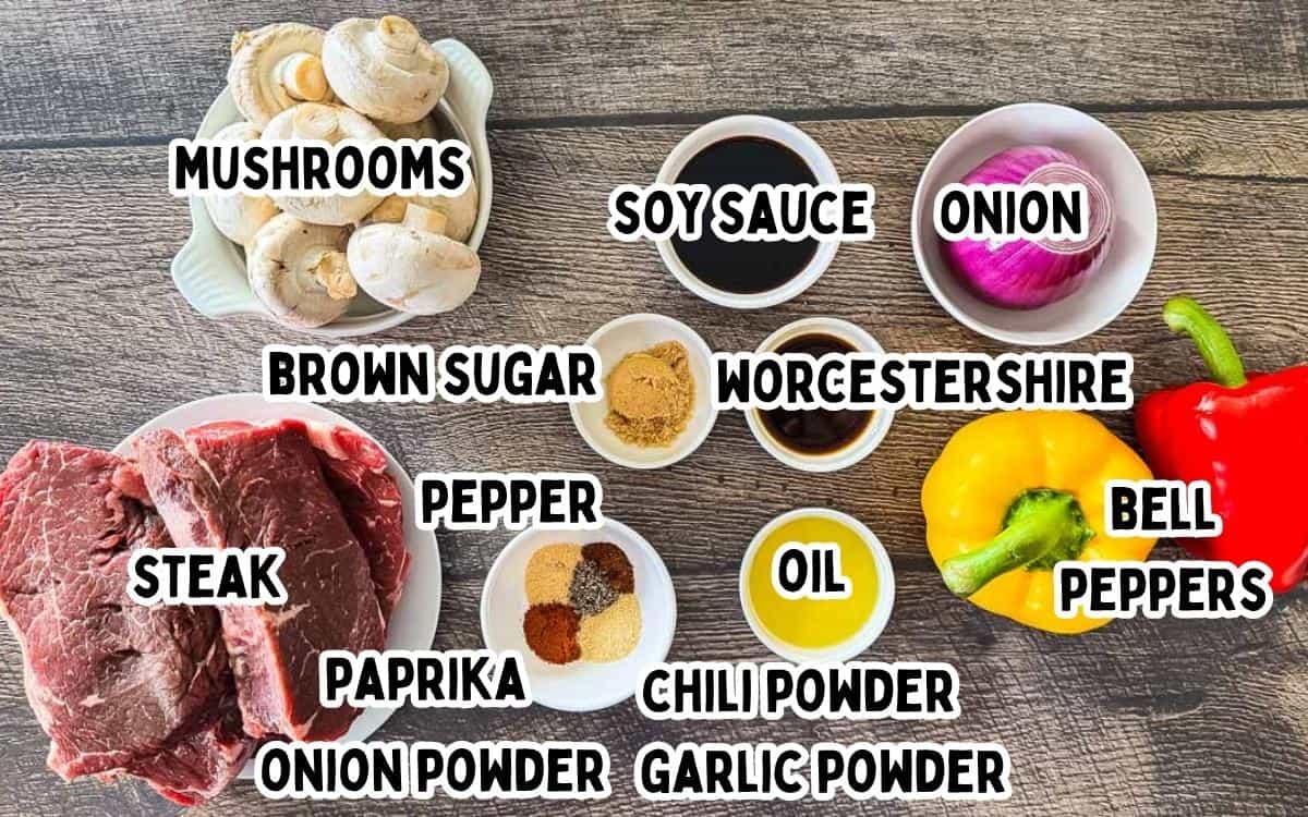 Ingredients for smoked steak kabobs including top sirloin steak, mushrooms, bell peppers, red onion, soy sauce, Worcestershire sauce, brown sugar, oil, and seasonings on a wooden table.