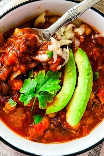 Smoked brisket chili topped with avocado, shredded cheese, and onions in a white bowl with a spoon lifting a hearty bite.
