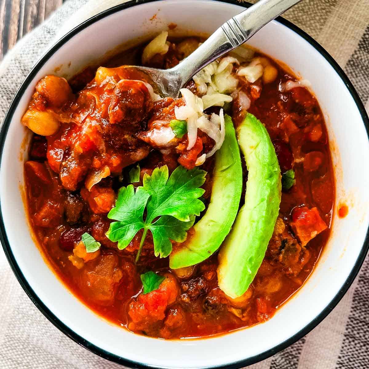 Smoked brisket chili topped with avocado, shredded cheese, and onions in a white bowl with a spoon lifting a hearty bite.