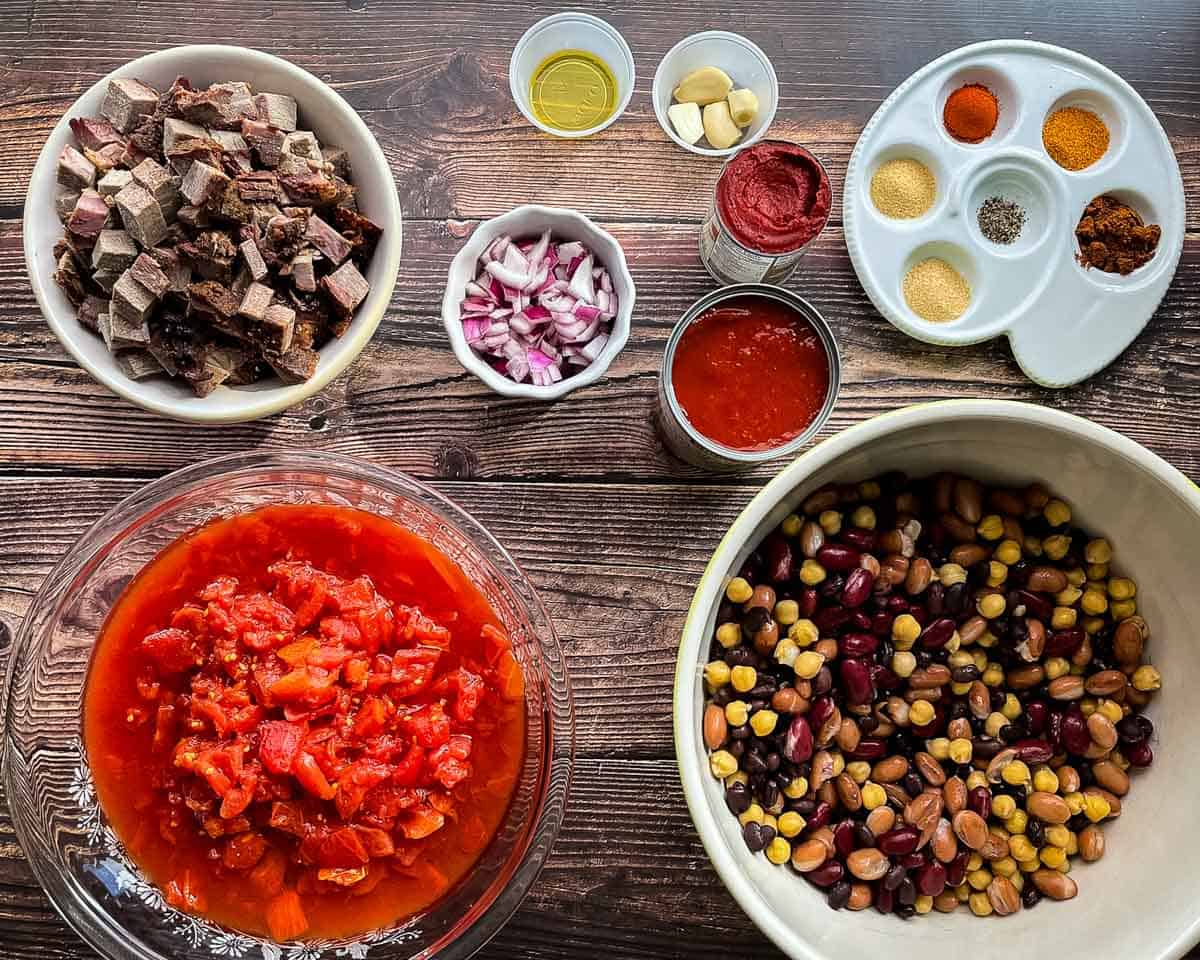 Ingredients for smoked brisket chili including leftover smoked brisket, mixed beans, tomatoes, tomato paste, red onion, garlic, and spices on a wooden table.