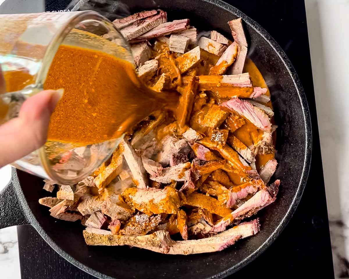 Taco seasoning mixed with water being poured over sliced smoked brisket in a cast iron skillet.