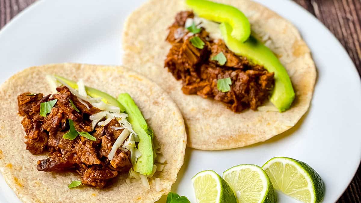 Smoked brisket tacos topped with avocado slices, shredded cheese, and cilantro on a white plate.