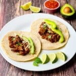 Two smoked brisket tacos topped with avocado slices, shredded cheese, and fresh cilantro on a white plate with lime wedges.