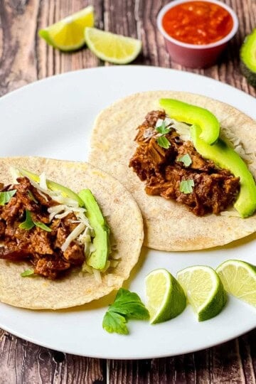 Two smoked brisket tacos topped with avocado slices, shredded cheese, and fresh cilantro on a white plate with lime wedges.