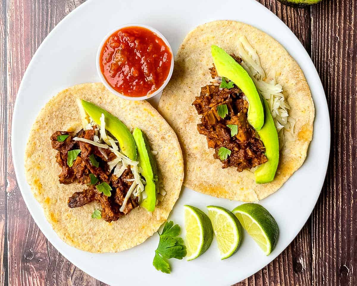 Smoked brisket tacos topped with avocado slices, shredded cheese, and cilantro on a white plate with salsa.