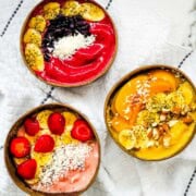 Three thick smoothie bowls topped with strawberries, bananas, blueberries, peaches, coconut, chia seeds, and almonds.
