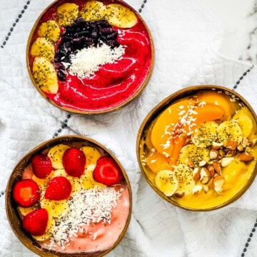 Three thick smoothie bowls topped with strawberries, bananas, blueberries, peaches, coconut, chia seeds, and almonds.