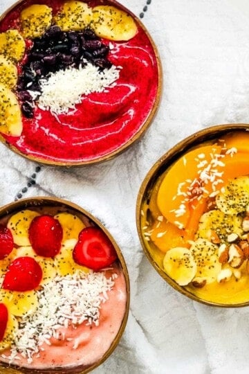 Three thick smoothie bowls topped with strawberries, bananas, blueberries, peaches, coconut, chia seeds, and almonds.