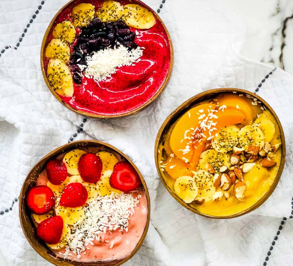 Three thick smoothie bowls topped with strawberries, bananas, mango, coconut, chia seeds, and almonds.