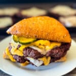 Double venison smash burger on a toasted bun topped with melted cheese, burger sauce, and sliced pickles, served on a white plate with smashed venison patties on a serving platter in the background.