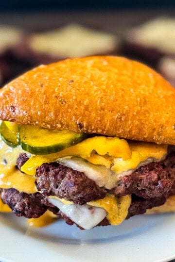 Double venison smash burger on a toasted bun topped with melted cheese, burger sauce, and sliced pickles, served on a white plate with smashed venison patties on a serving platter in the background.